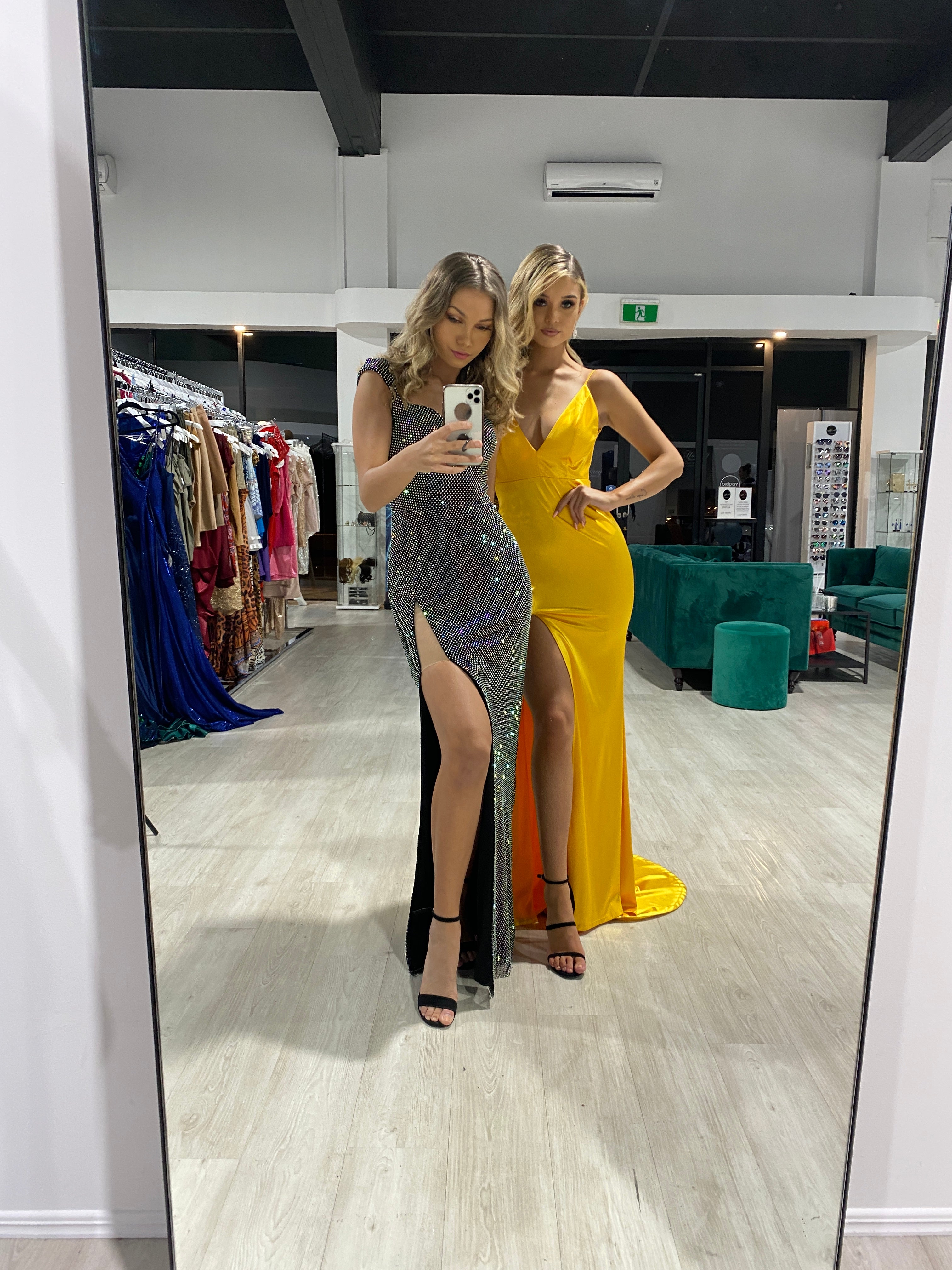 Two women in a boutique, one wearing a glittery black dress with a side slit, the other in a yellow evening gown, posing in front of a mirror.