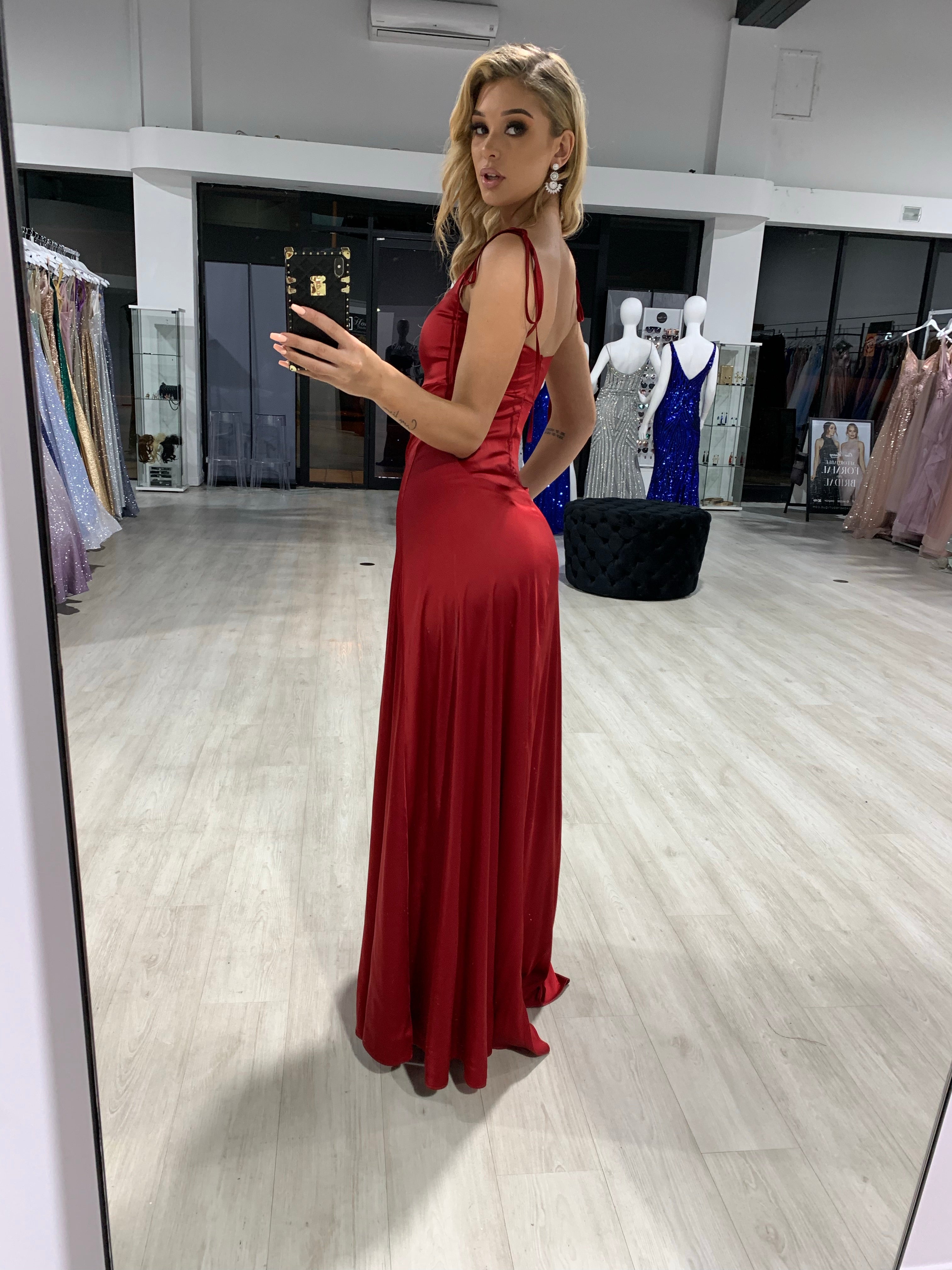 Woman wearing Honey Couture XENIA Burgundy Tie Up Formal Bridesmaid Dress in store setting, showcasing evening gown with tie-up straps.