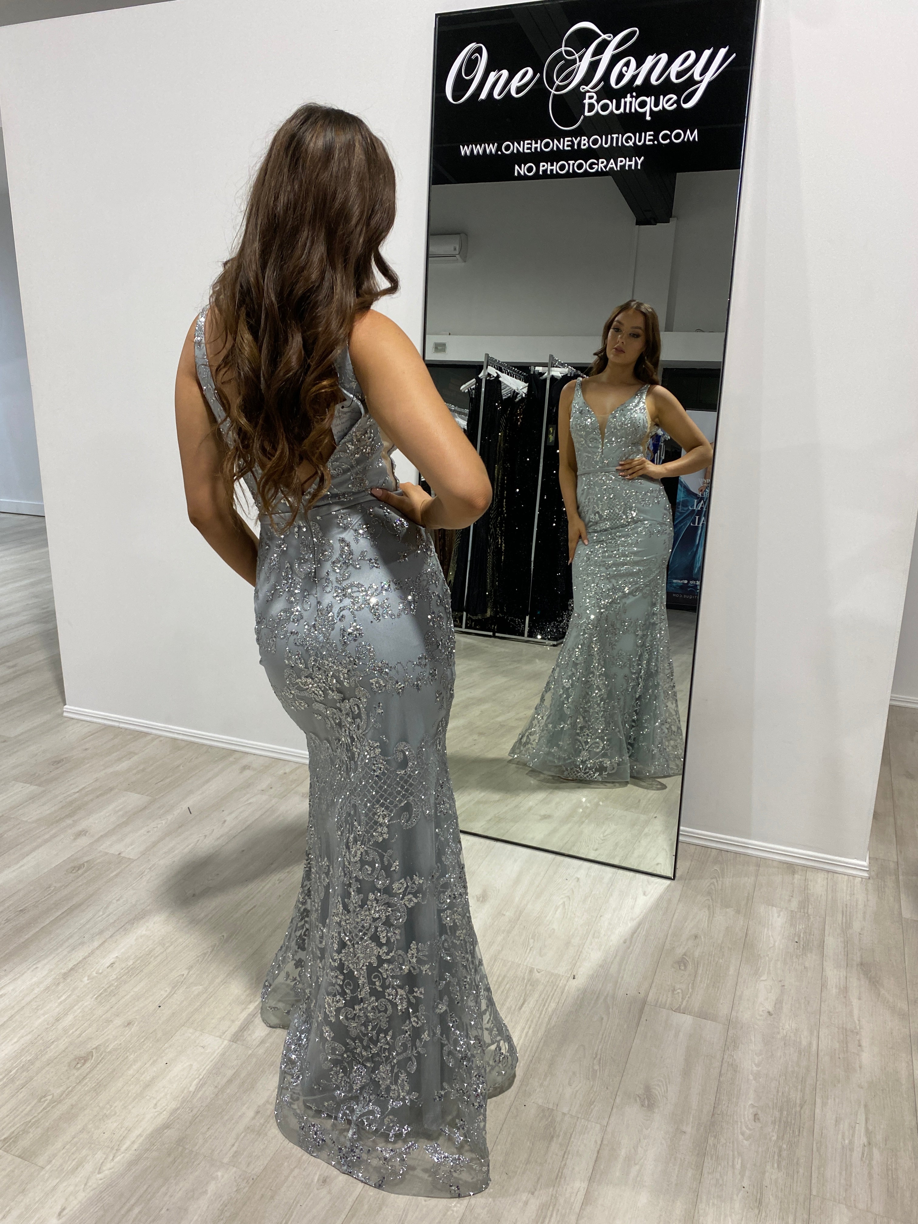 Woman wearing Honey Couture CAROLINE Silver Mesh Glitter Formal Gown, an Australian Designer prom dress, admiring her reflection in a mirror.