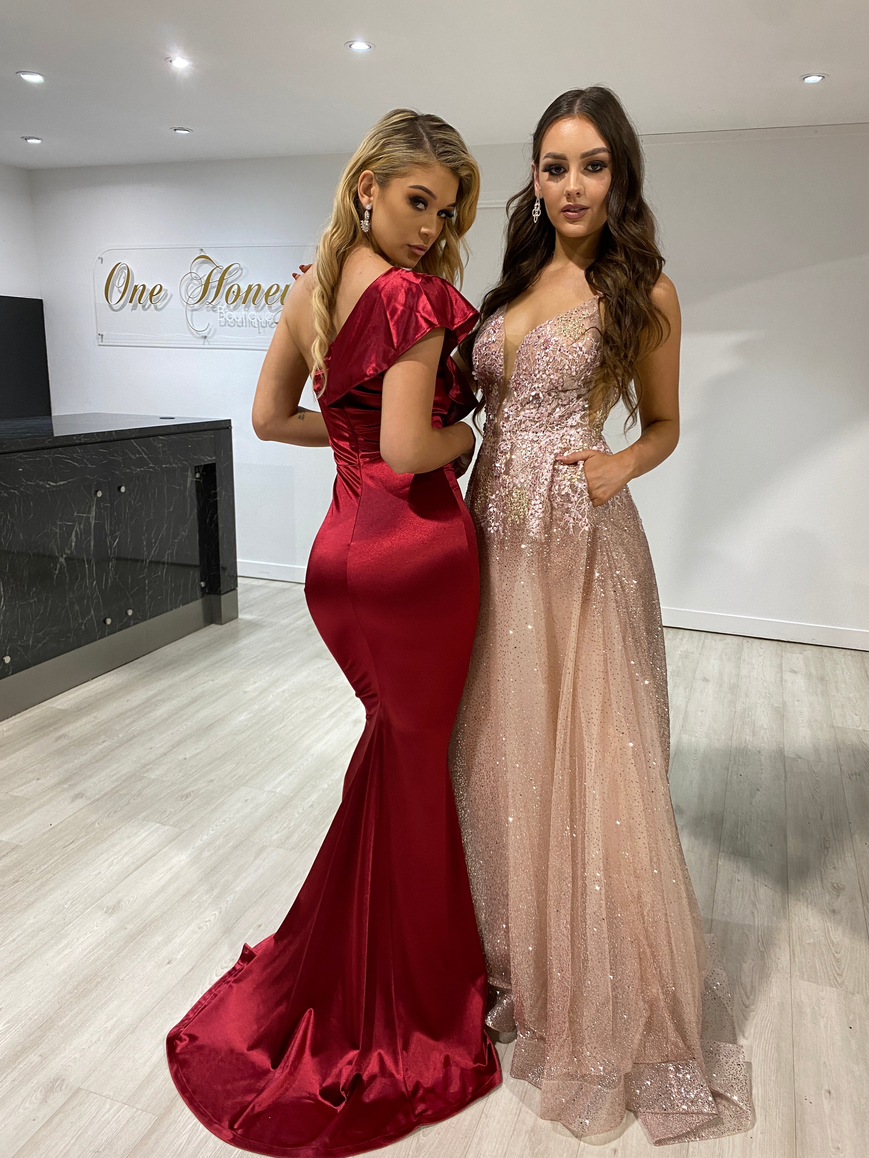 Two women wearing elegant formal dresses, one in red satin and the other in a Honey Couture SIERRA rose gold and pink glitter gown, posing indoors.