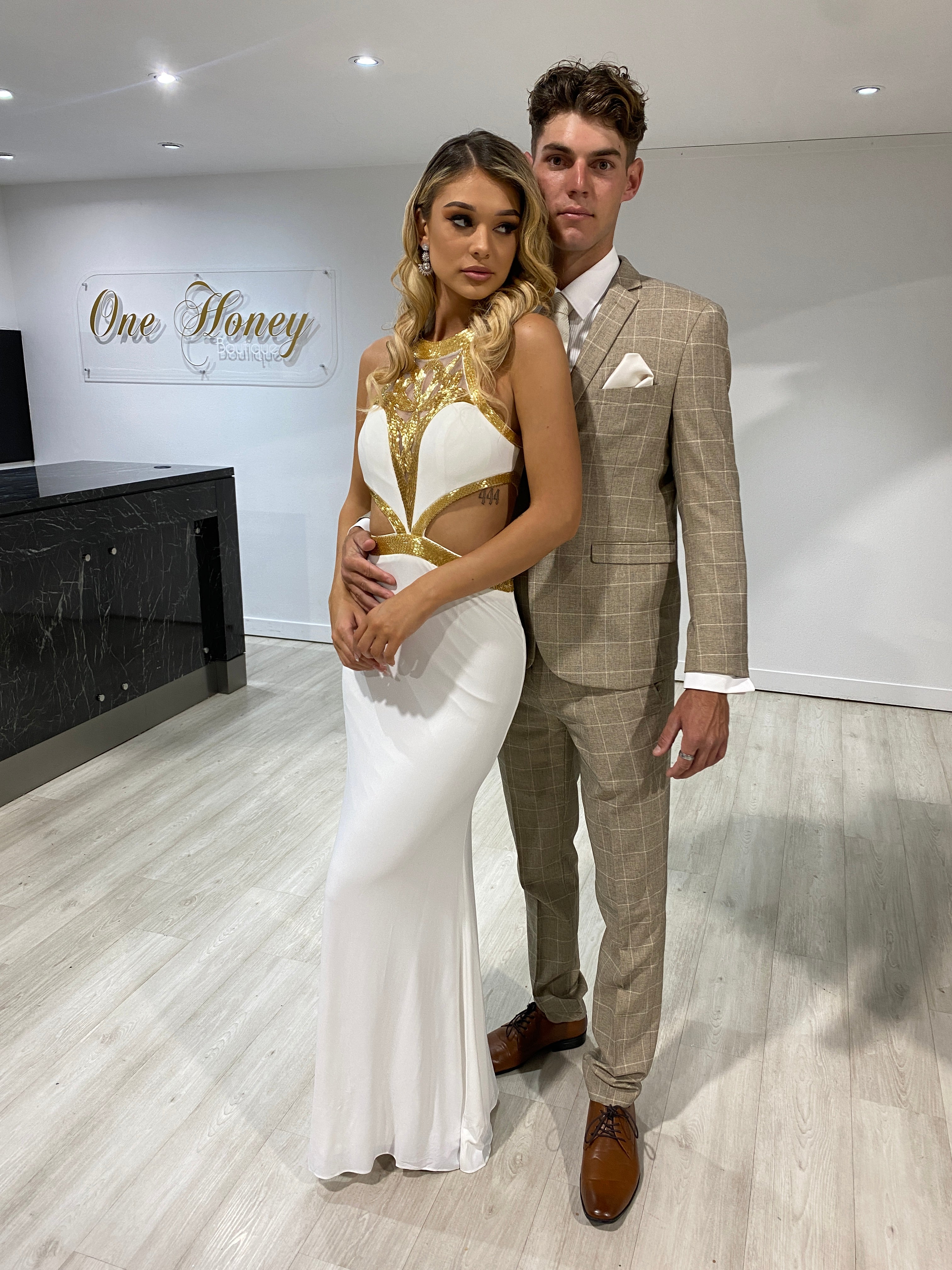 Couple dressed in elegant formal attire, with the woman in a white gown and the man in a beige suit, standing in a modern lobby.