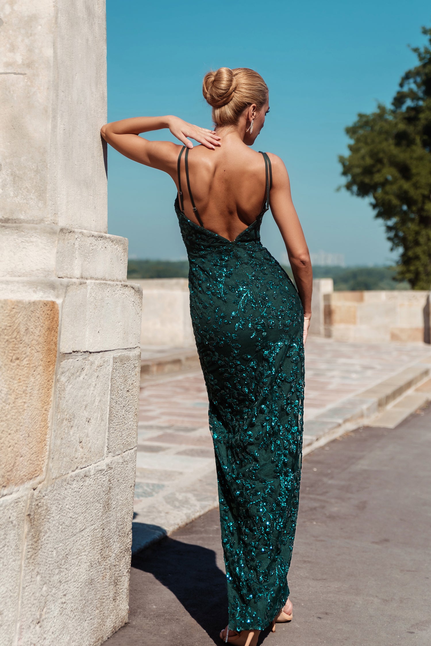 Back view of a woman in a sparkling emerald green evening gown with thin straps, standing by a stone wall.