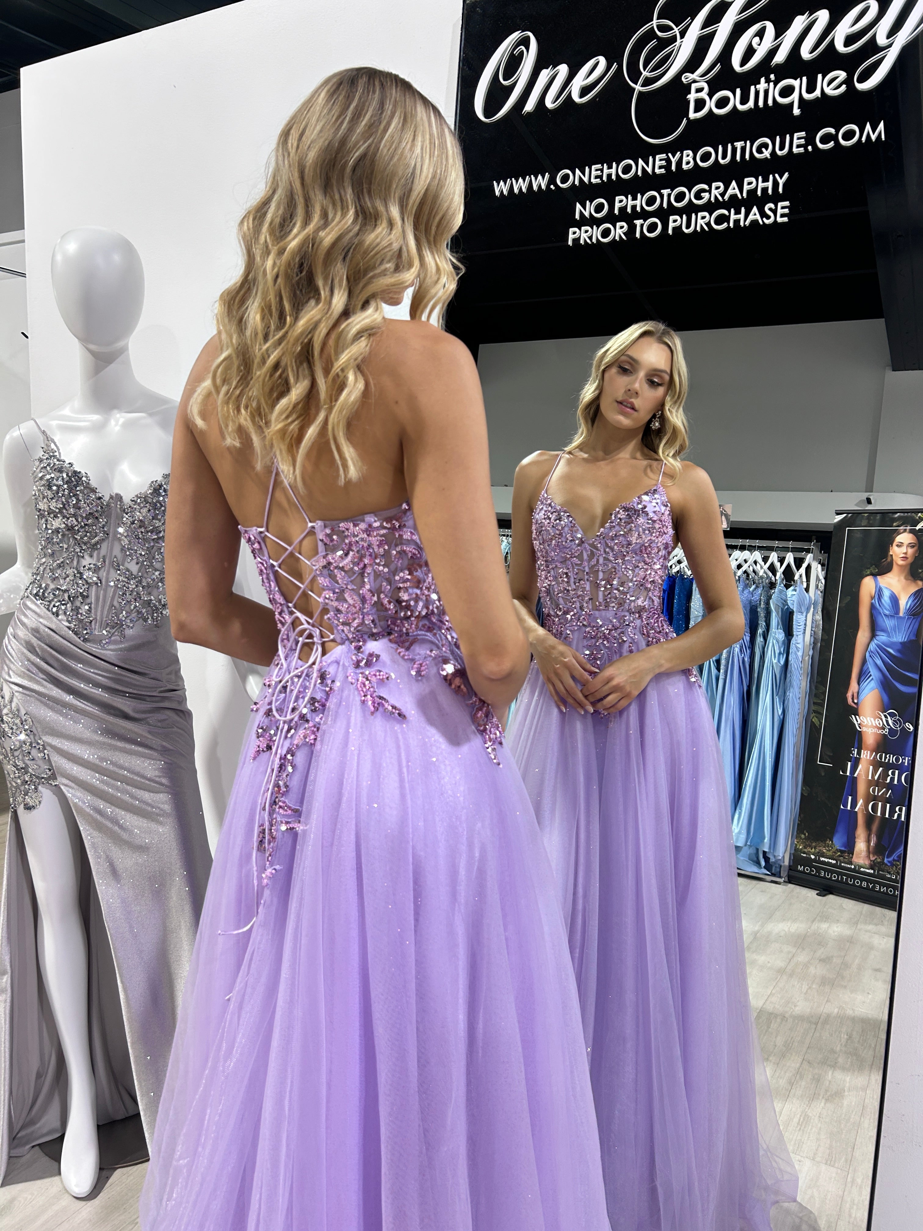 Woman admiring herself in a mirror wearing a purple Honey Couture NOVINE sequin and glitter mesh tulle prom dress at a boutique.