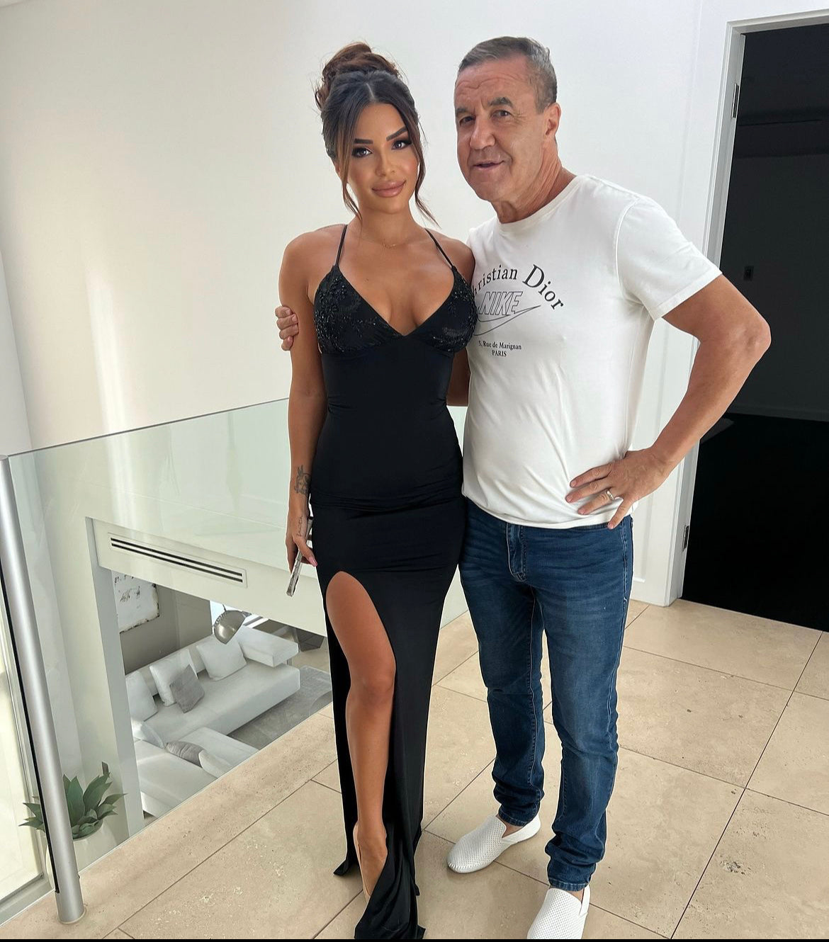 Woman in black evening dress with lace detail and side slit, standing next to a man in casual attire indoors.